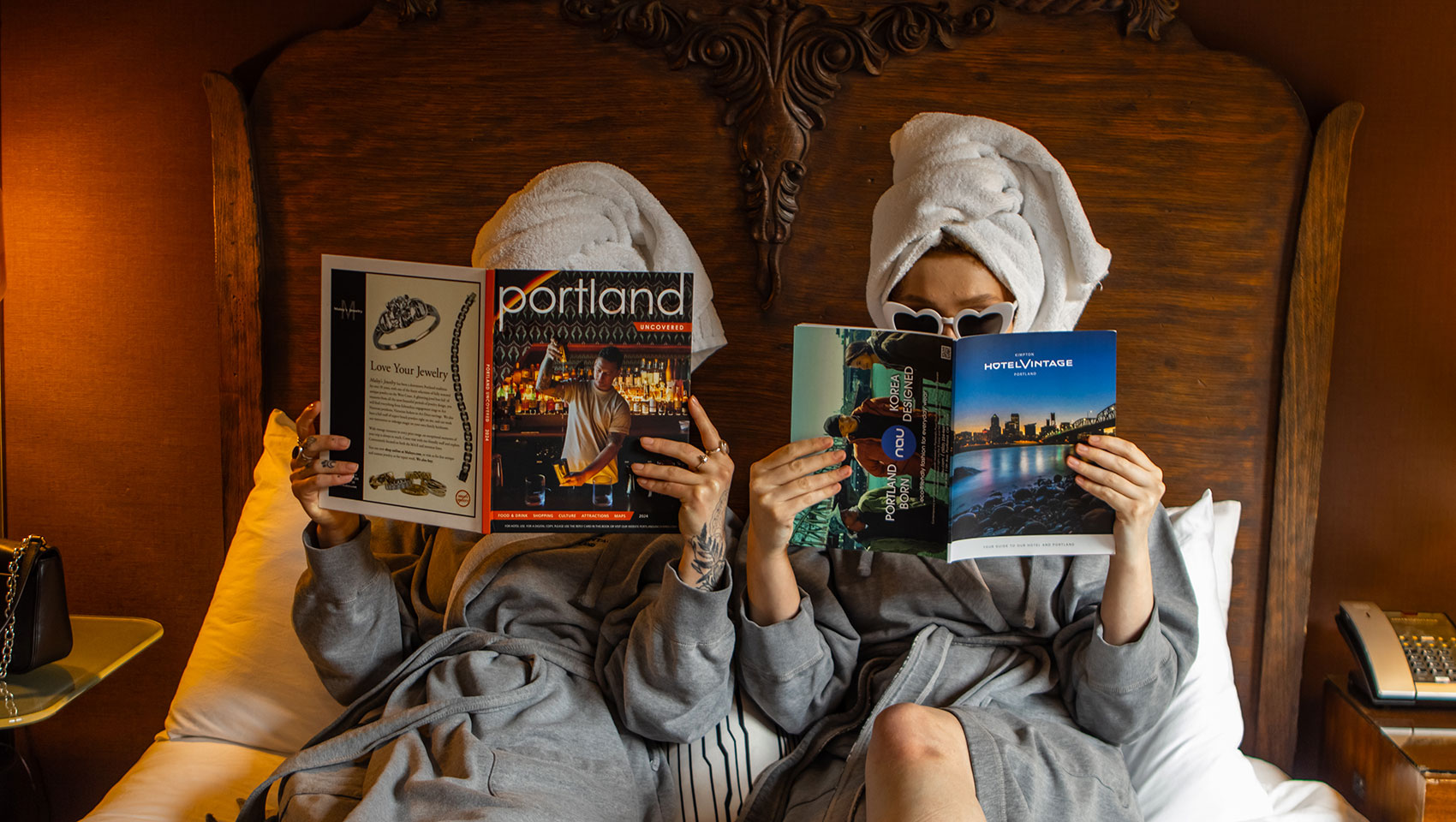 Two women in robes with towels around their hair sitting on a bed while reading magazines